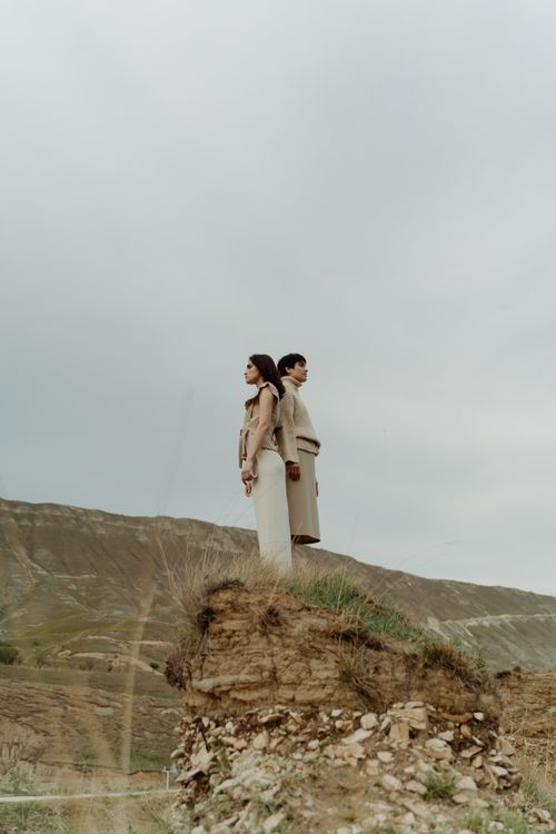 Free Two adults in neutral attire standing back to back on a rocky hill with cloudy sky backdrop. Stock Photo