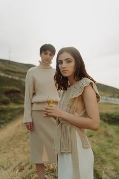 Two women in knitted outfits posing outdoors with flowers, showcasing modern fashion.