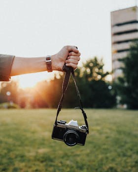 A hand holding a vintage camera with a grassy background during sunset outdoors.