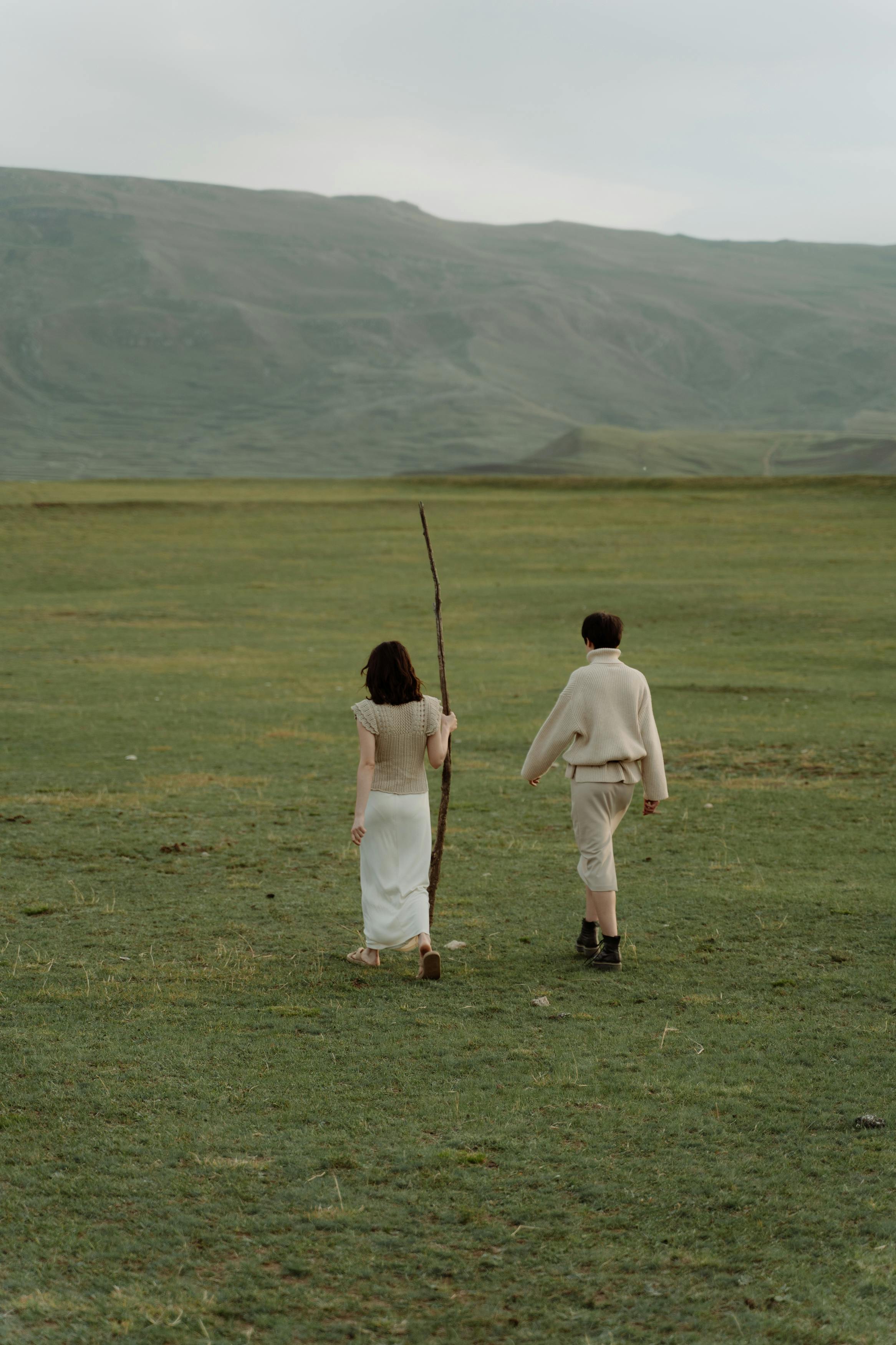 A couple walking on a vast grassland, one holding a stick, against rolling hills.