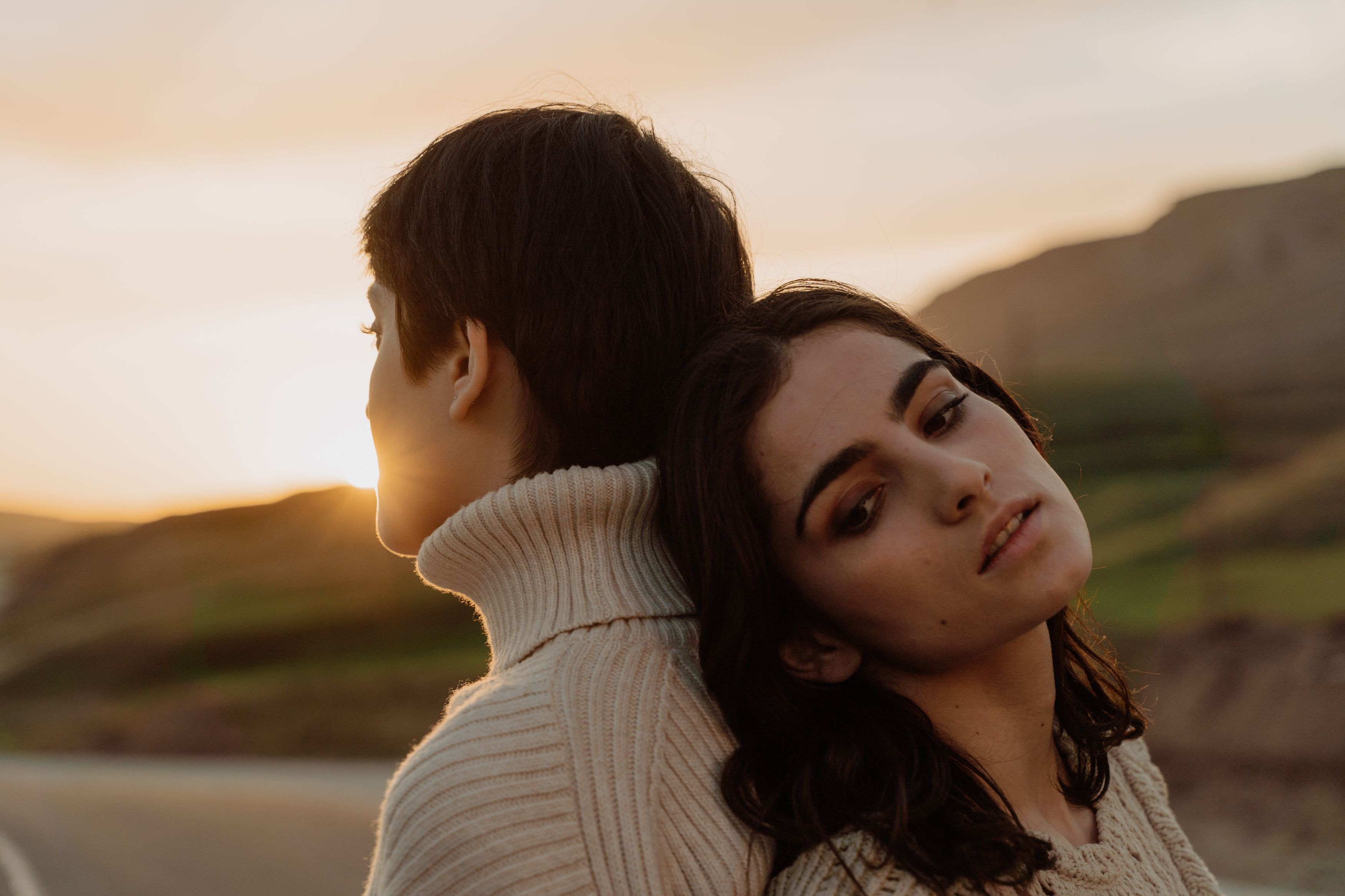 Two women in warm sweaters pose back-to-back at sunset, creating a serene and intimate atmosphere.