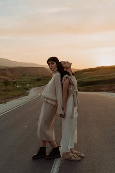Two stylish women in fashionable outfits pose back-to-back on a scenic country road during sunset.