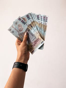 Hand holding 100 banknotes in a fan shape on a neutral background.