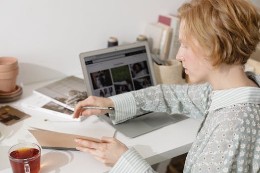 A woman in a floral top works at a white desk with a laptop, notebook, and cup of tea.