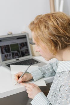 Adult woman writing notes at home workspace with a laptop in a cozy setting.