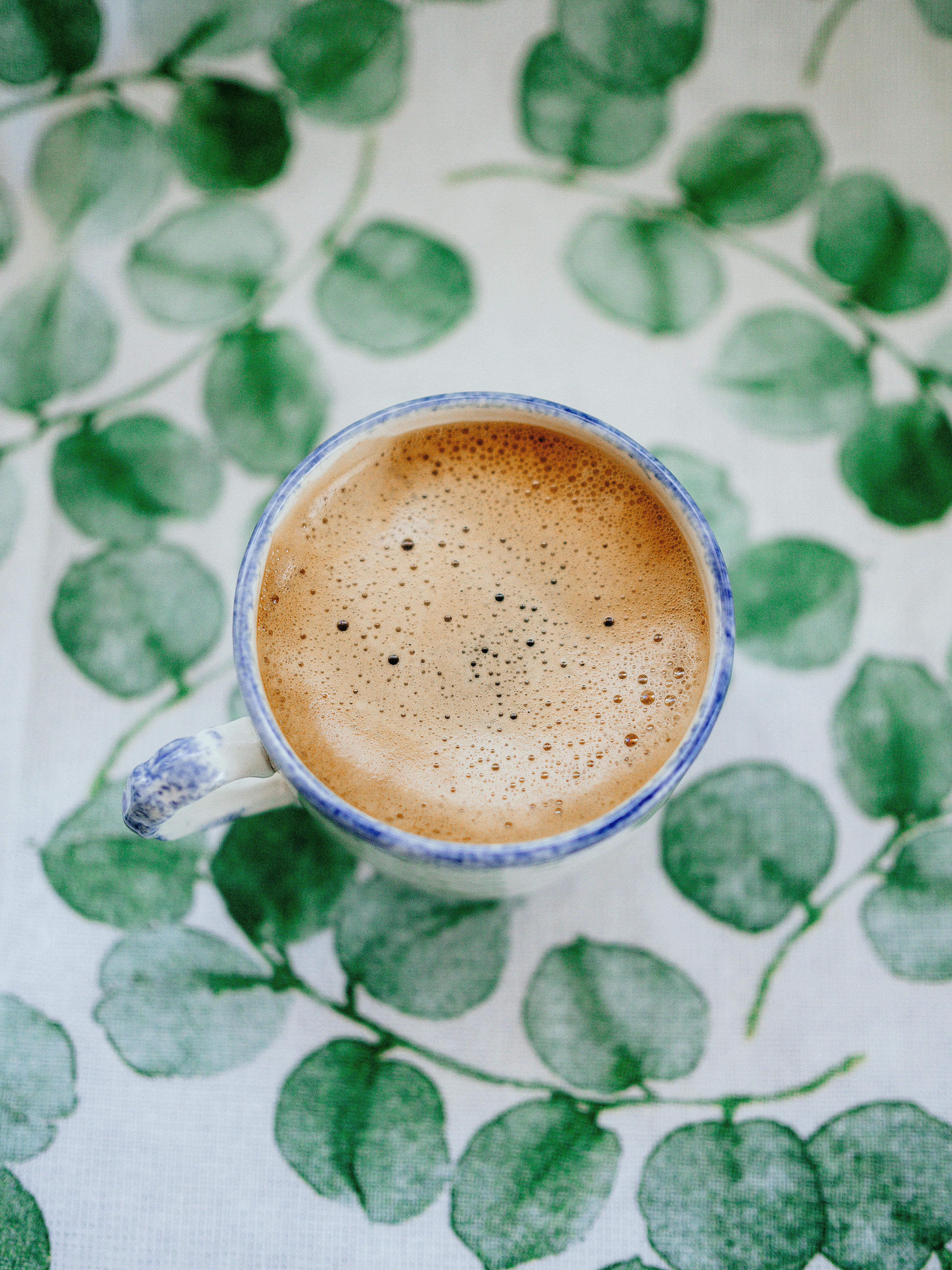 A hot cup of coffee with frothy texture sits on a green leafy patterned tablecloth, captured from above.