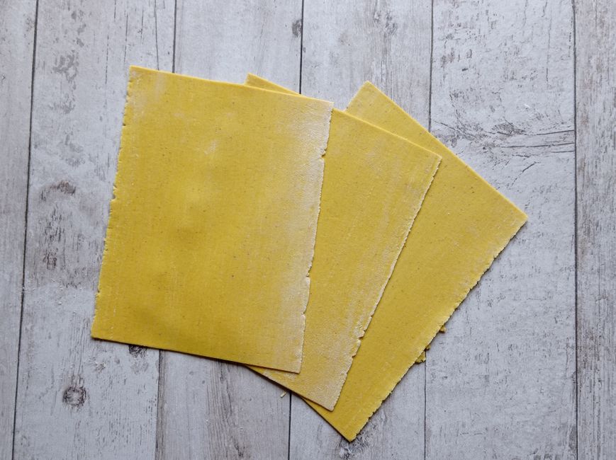 A stack of flat, wide, ridge-edged dry lasagna noodles next to a bowl of fresh pasta sheets cut into rectangles.