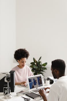 Two diverse colleagues working together on laptops in a bright, modern office setting.