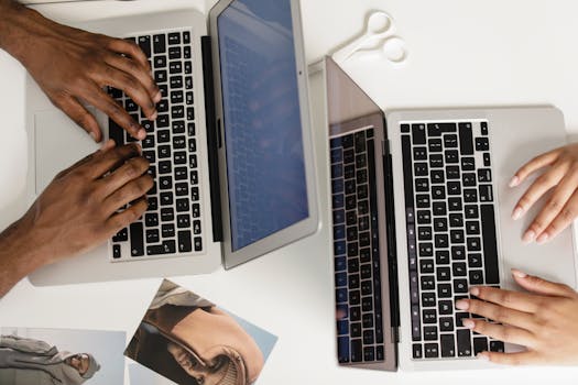 Two hands typing on laptops on a white desk, viewed from above, fostering teamwork.