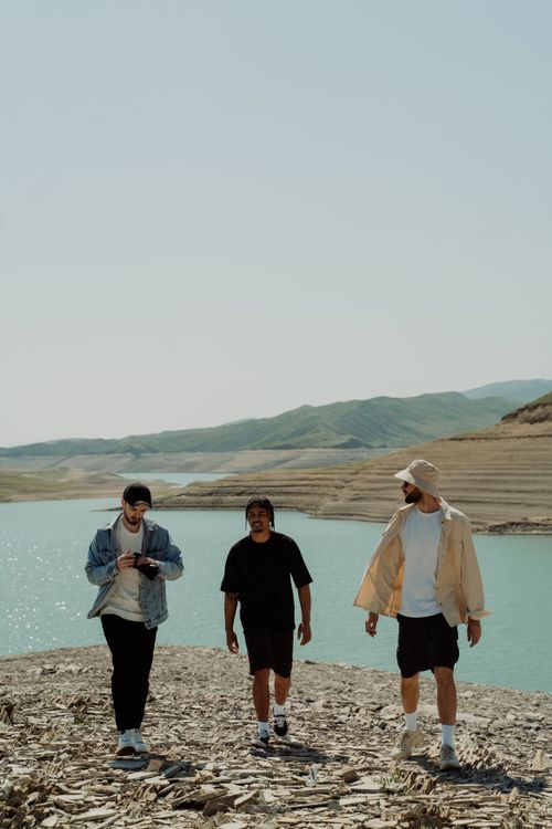 Free Three men enjoying a walk by a picturesque lake with mountain views on a clear day. Stock Photo