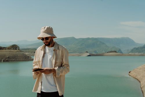 Free A stylish man with a bucket hat stands by a serene lake, enjoying the picturesque landscape. Stock Photo