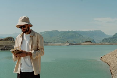 Free A man wearing a bucket hat and sunglasses looks at a book by a peaceful reservoir. Stock Photo