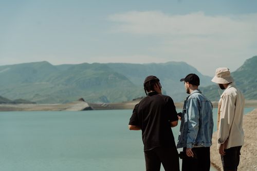 Free Three men in hats stand by a serene mountain lake, enjoying the scenic landscape under a clear sky. Stock Photo