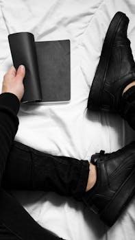 Overhead shot of person wearing black sneakers and pants, holding a notebook on white bedding.