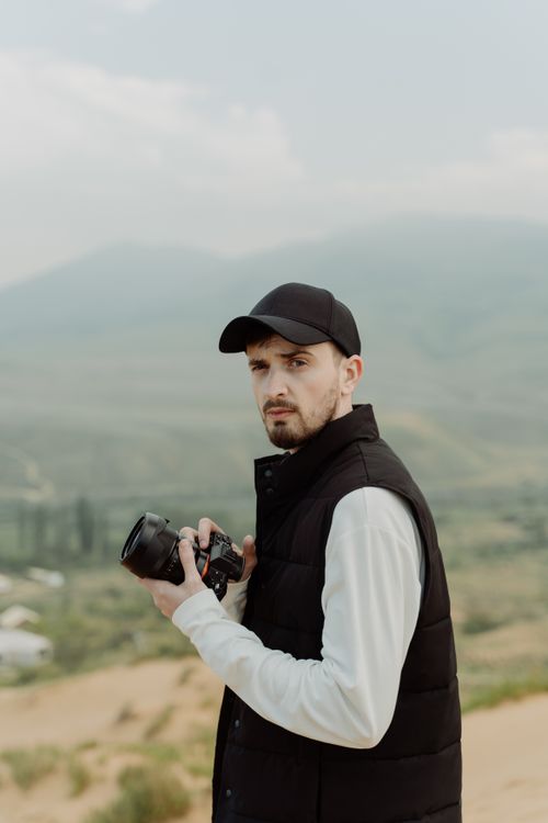 Free A photographer in a black vest with a DSLR camera captures a serene countryside scene. Stock Photo