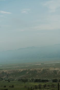 A tranquil misty view of expansive green fields and distant mountain ranges.