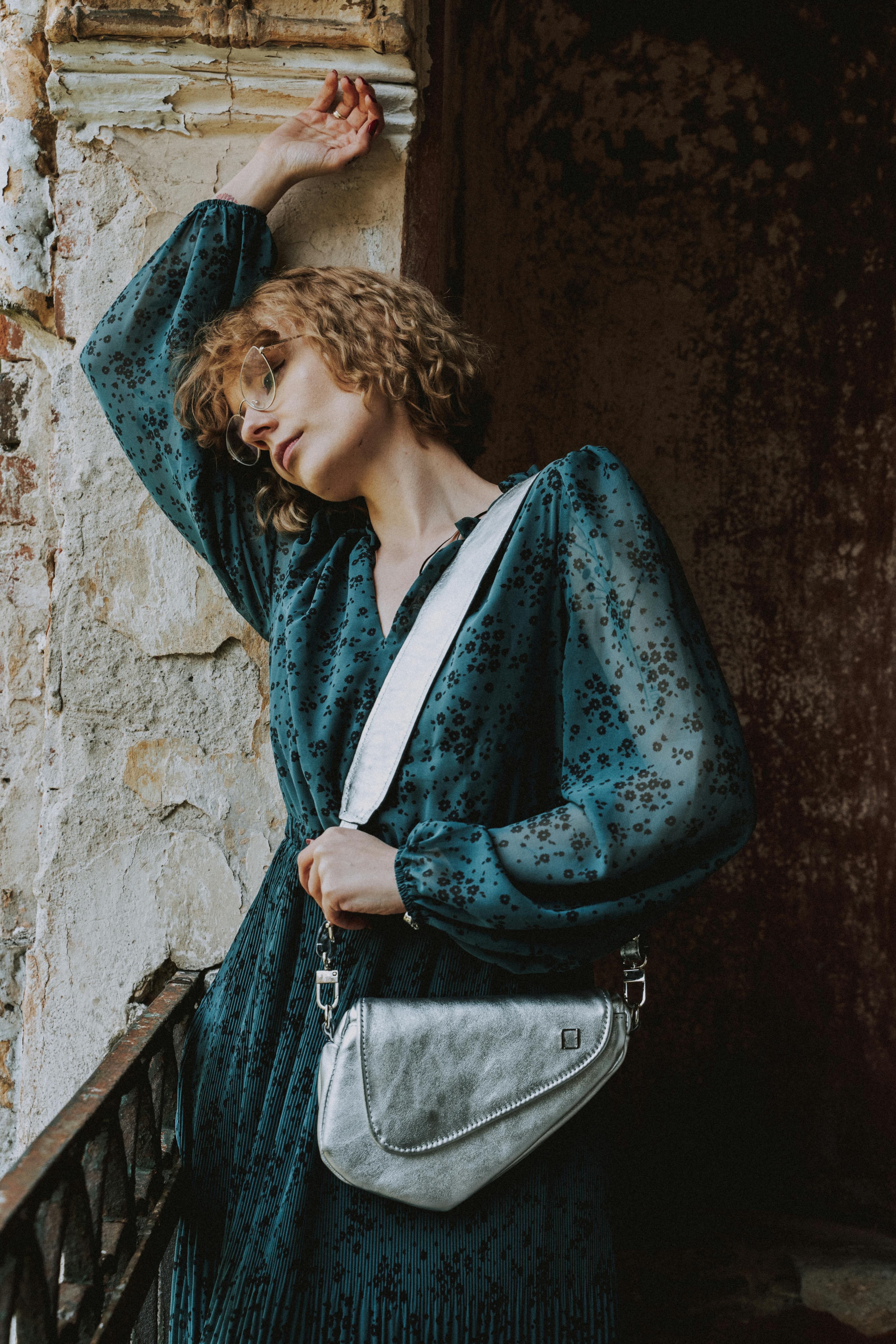 Elegant woman with short hair in a teal dress and eyeglasses, leaning thoughtfully against a rustic wall.