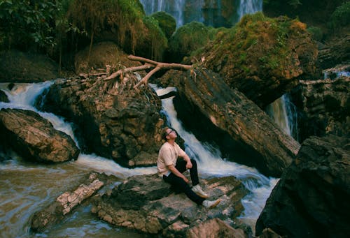 免费 男子坐在瀑布周围的灰色岩石上的照片 素材图片