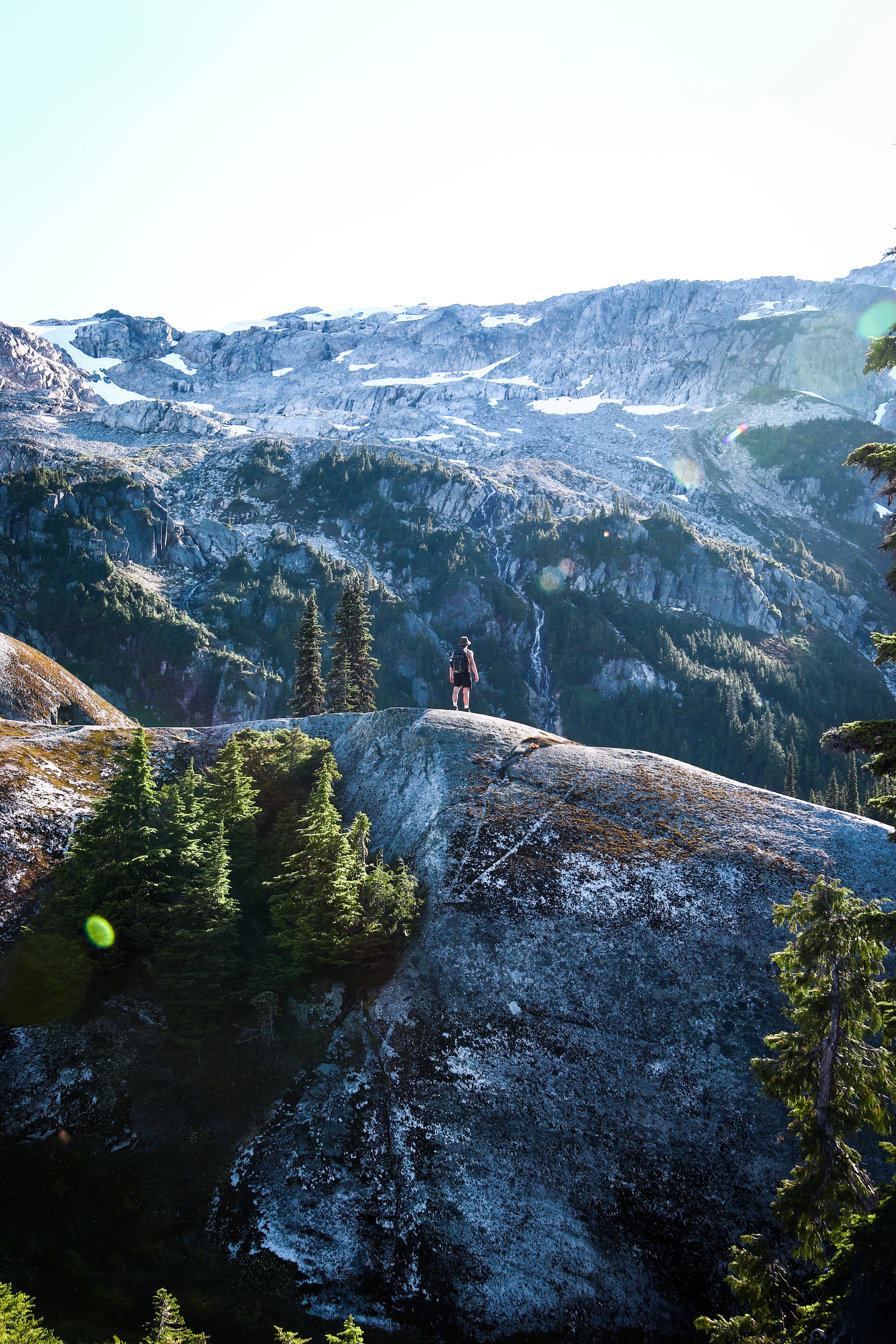 A lone hiker stands atop a rocky peak amidst lush trees with majestic mountains in Whistler, BC, Canada.