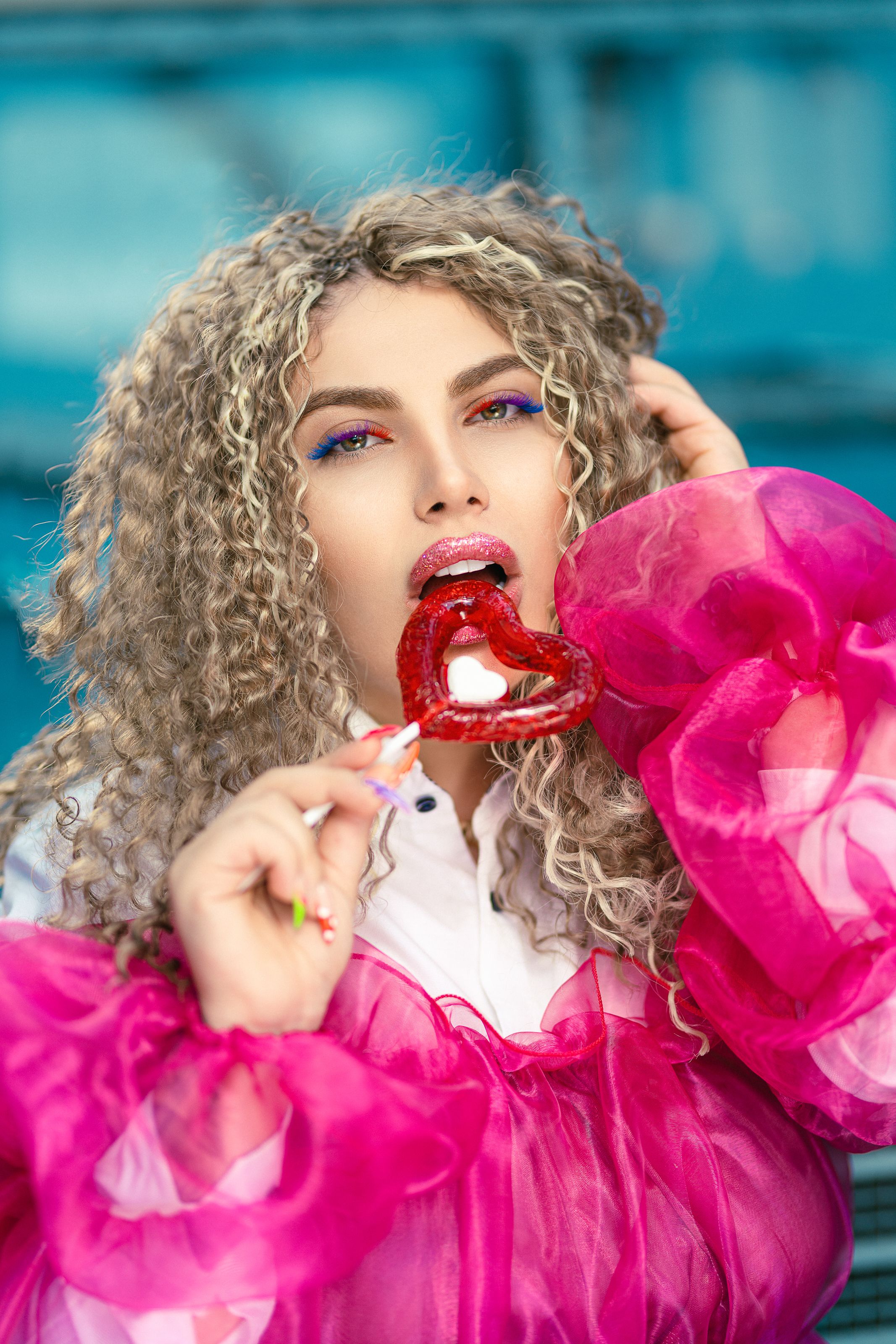 Curly-haired woman in vibrant attire savoring a heart-shaped lollipop outdoors.