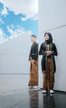 A couple in traditional clothing poses for a prewedding shoot, showcasing cultural attire against a modern architectural backdrop. - Fotografer Sidoarjo