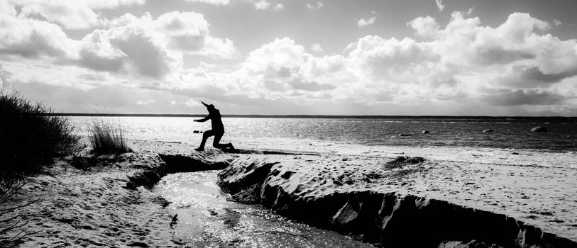 A stunning black and white photo capturing a silhouette jumping along a tranquil beach at sunset.
