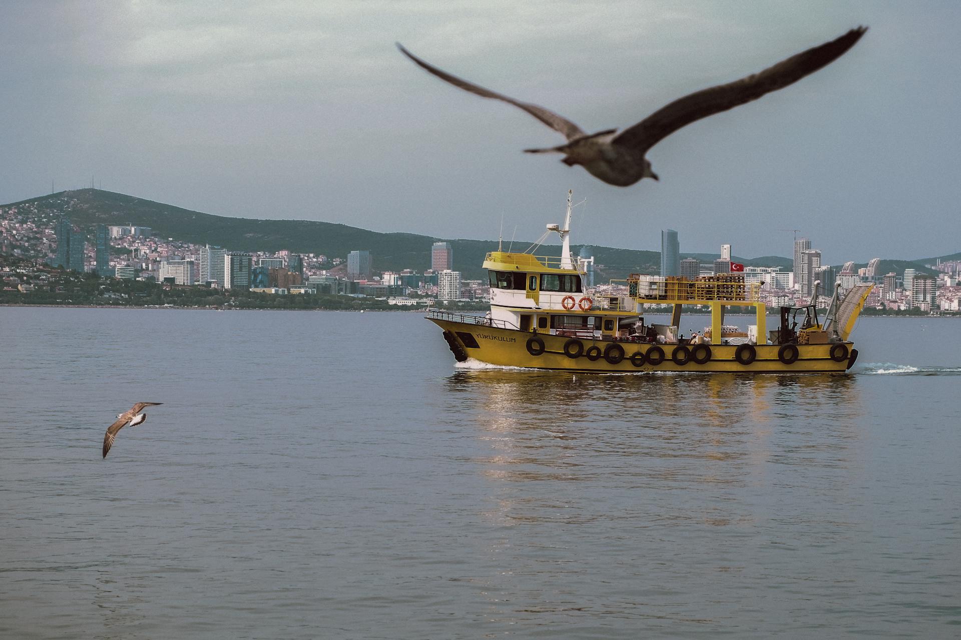 A yellow tugboat cruises on calm waters with city skyline and seagulls in the background.
