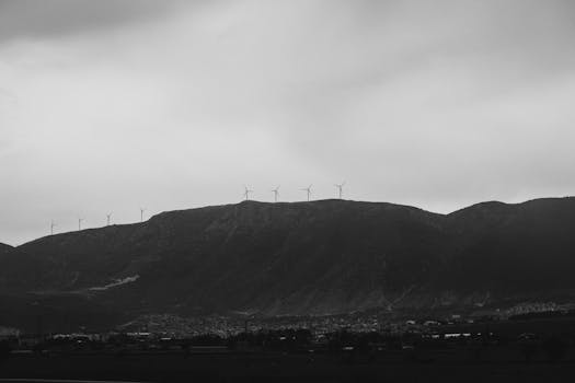 A grayscale image of wind turbines spread across a mountainous landscape under a cloudy sky.
