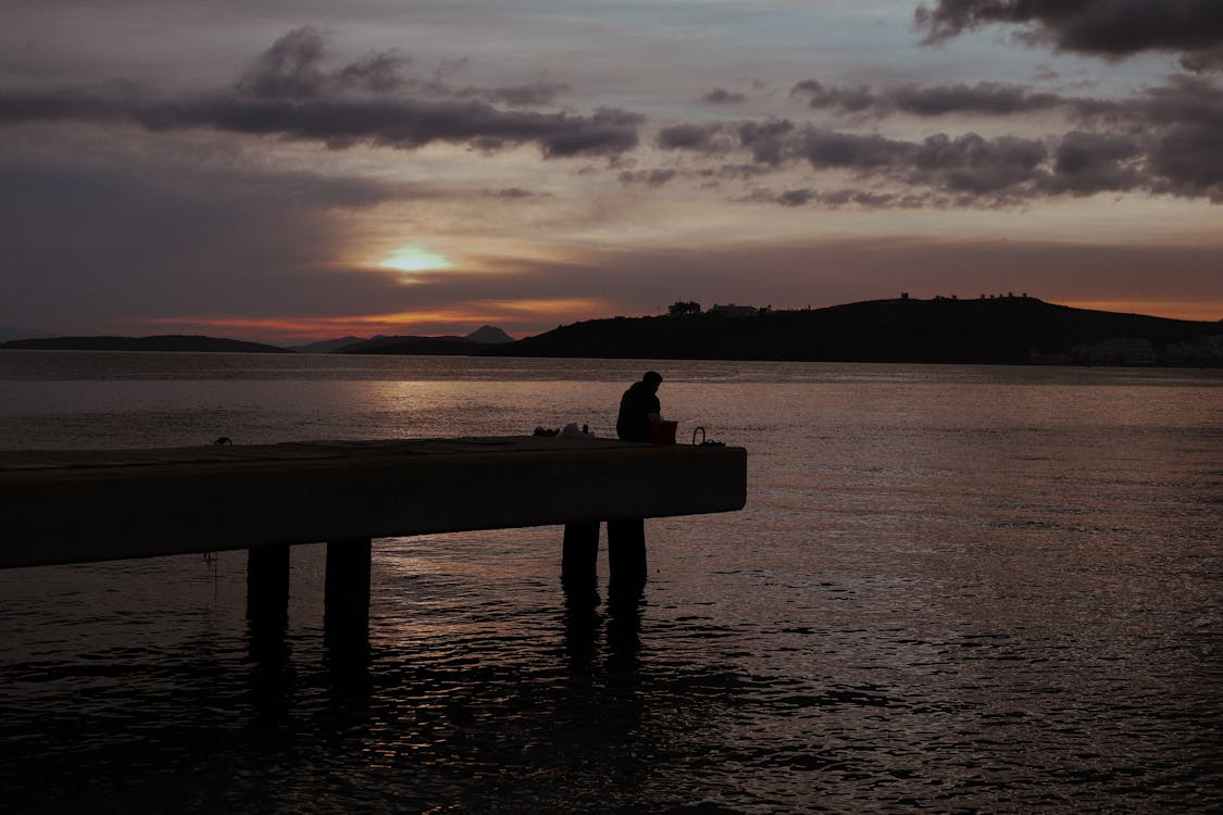 Silhouette of a Person Sitting on Concrete Dock during Sunset