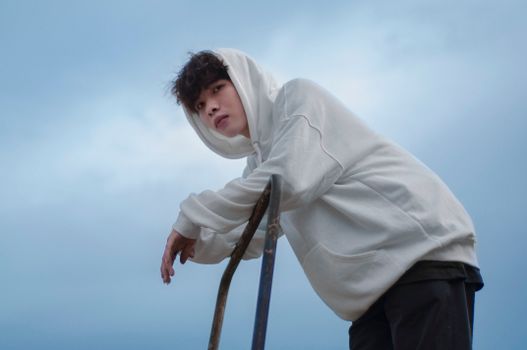 Portrait of a young man in a white hoodie leaning on a metal railing under a blue sky.