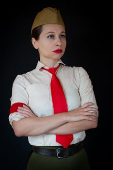 Woman in a red tie and cap poses with arms crossed against dark background.