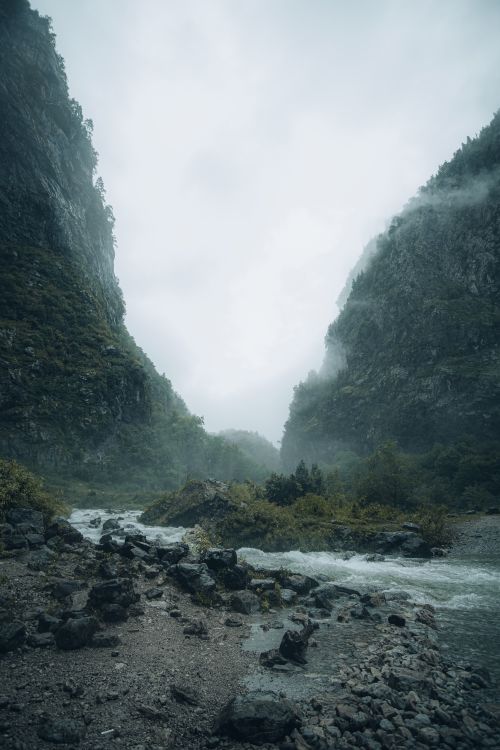 Free Serene misty mountain landscape featuring a flowing river surrounded by lush greenery and rugged rocky terrain. Stock Photo