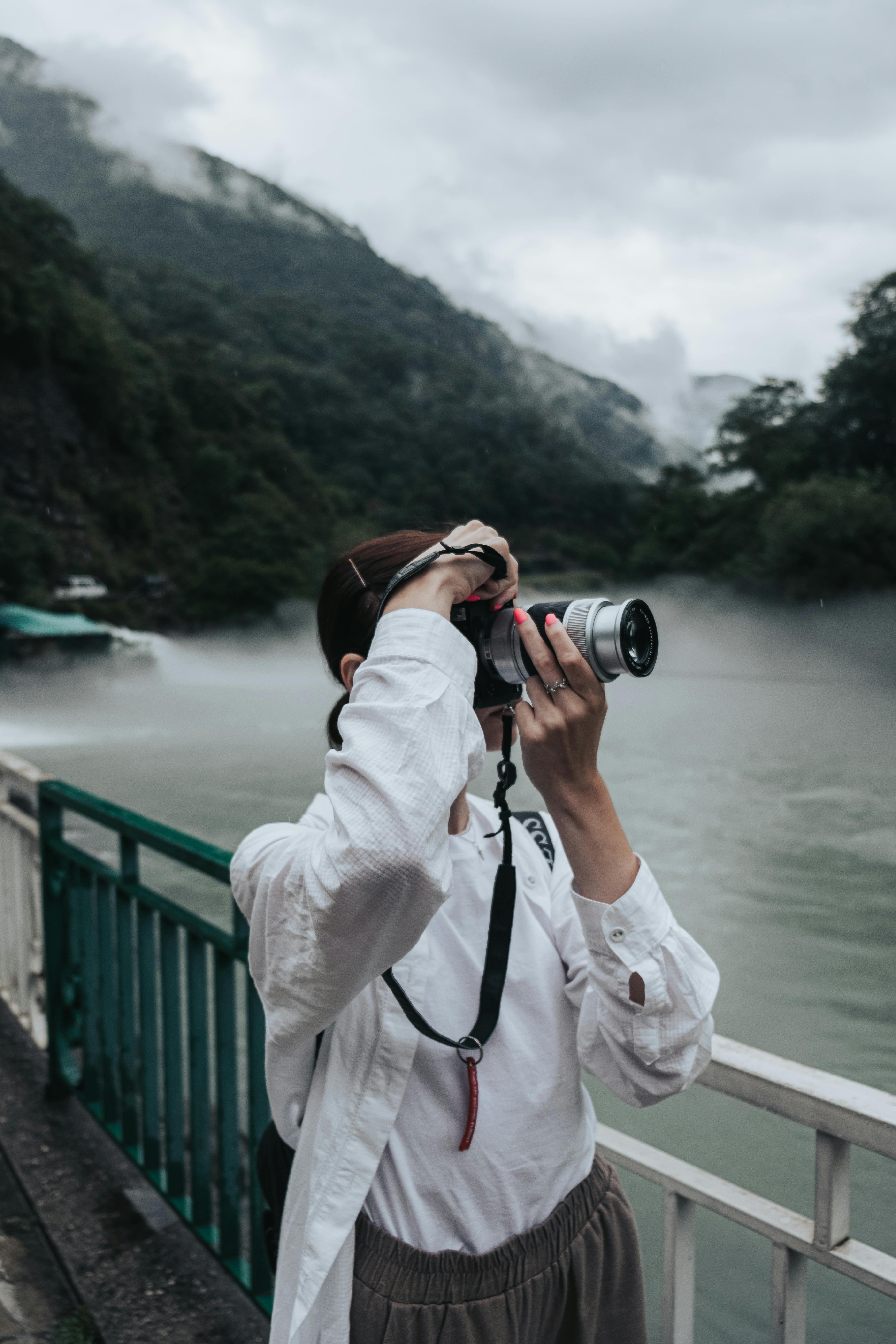 A woman stands on a bridge taking pictures of a foggy river and mountains.
