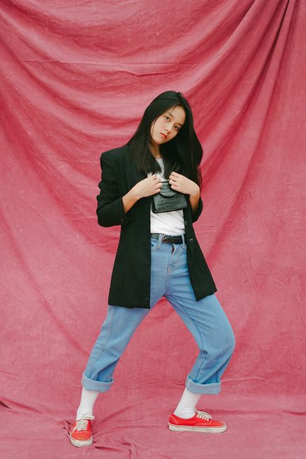 Stylish woman in black coat, blue jeans, and red sneakers posing confidently in a studio with a red draped backdrop.