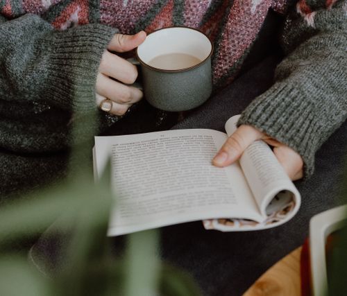 Free Person holding a coffee mug while reading a book indoors, exuding a warm and cozy atmosphere. Stock Photo