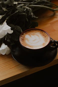 Elegant cappuccino with latte art next to white flowers on a wooden table, showcasing calm lifestyle moments.
