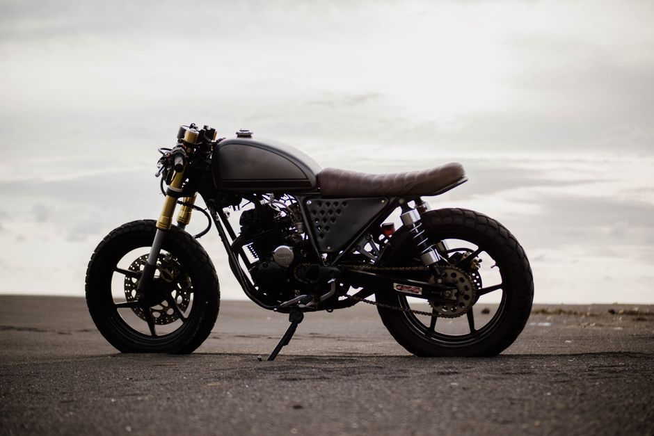 Side view of a vintage cafe racer motorcycle parked on an empty road under a cloudy sky.