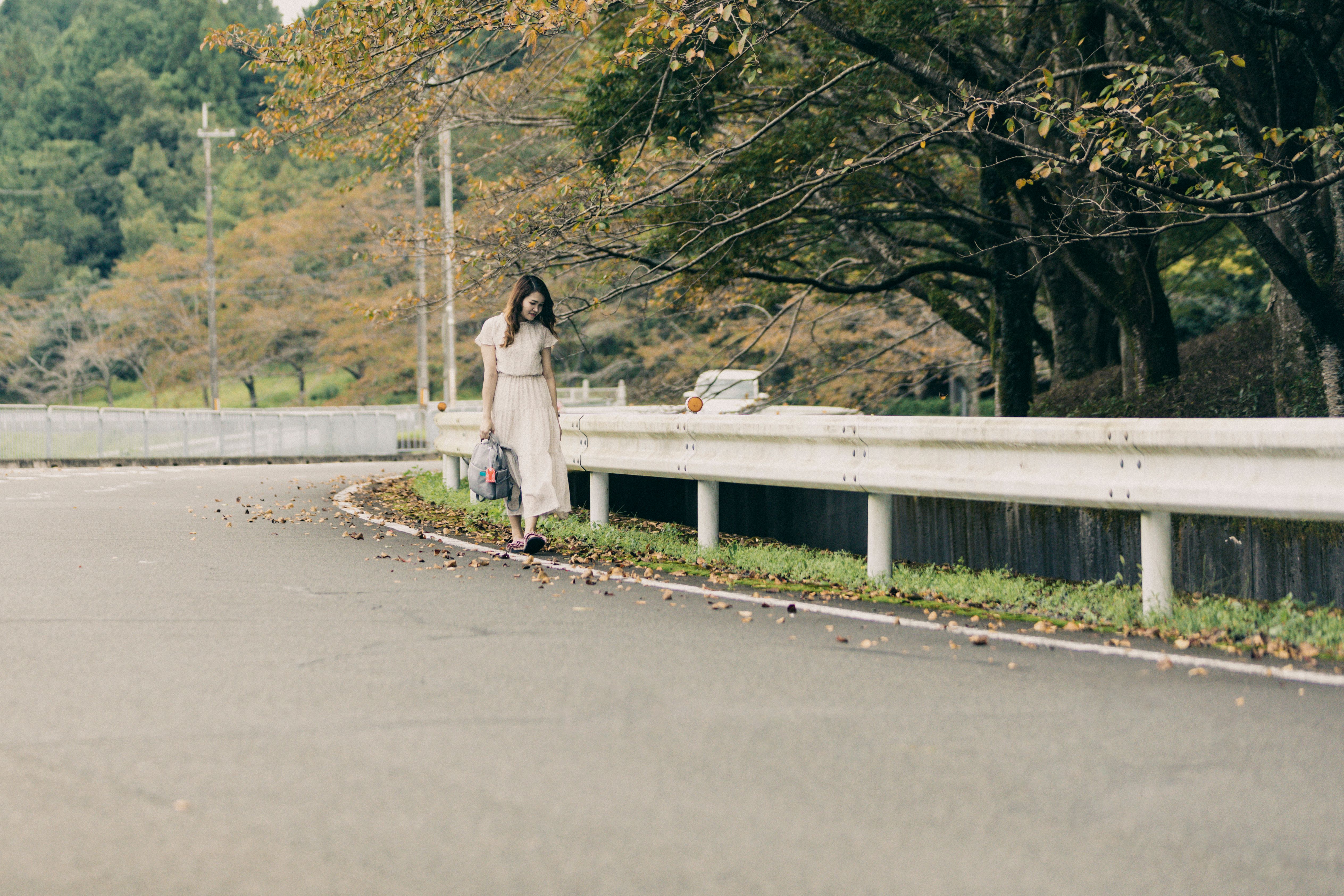 A woman in a white dress walks along a scenic countryside road lined with trees.