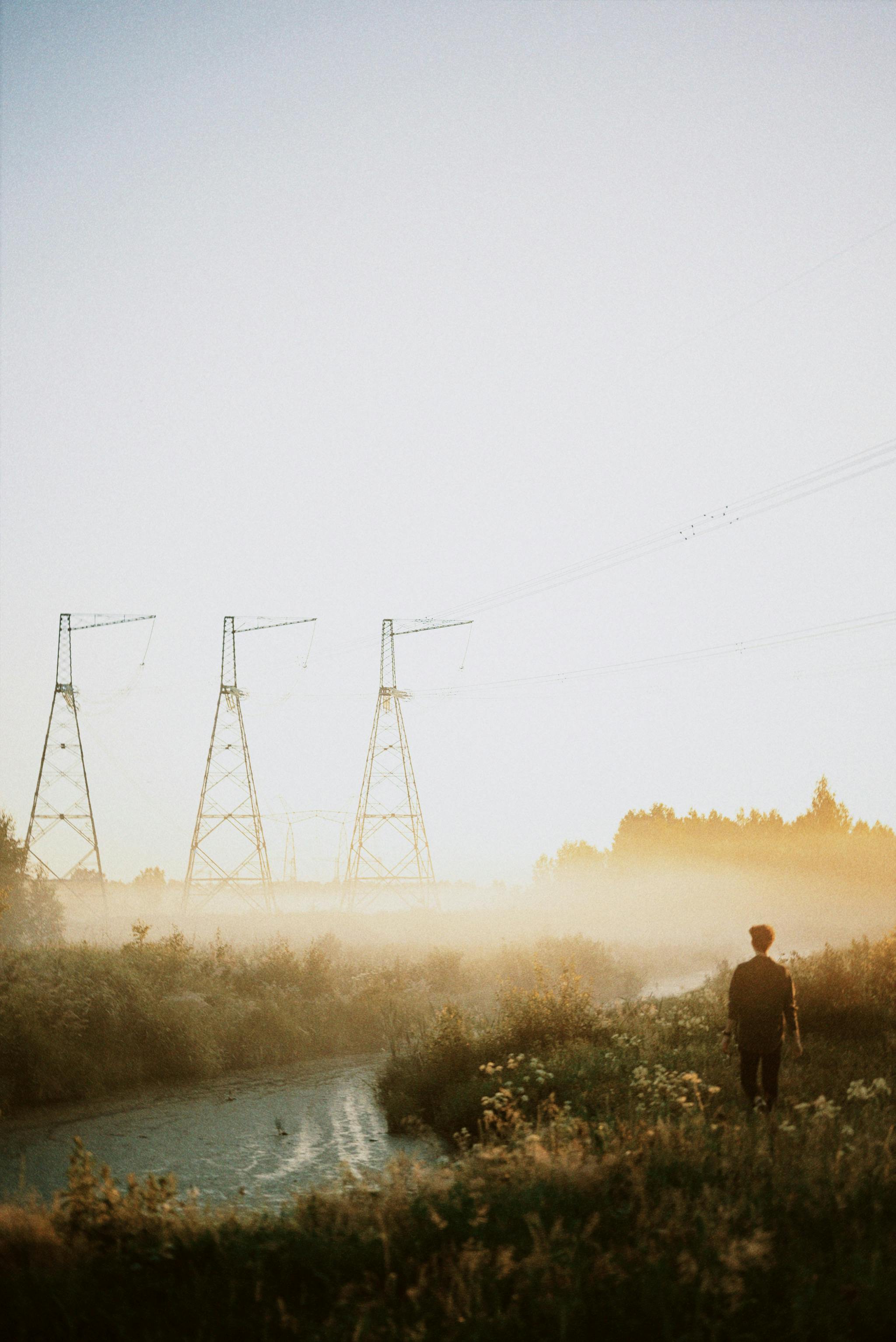 A solitary figure stands by a misty river with power lines in rural Russia.