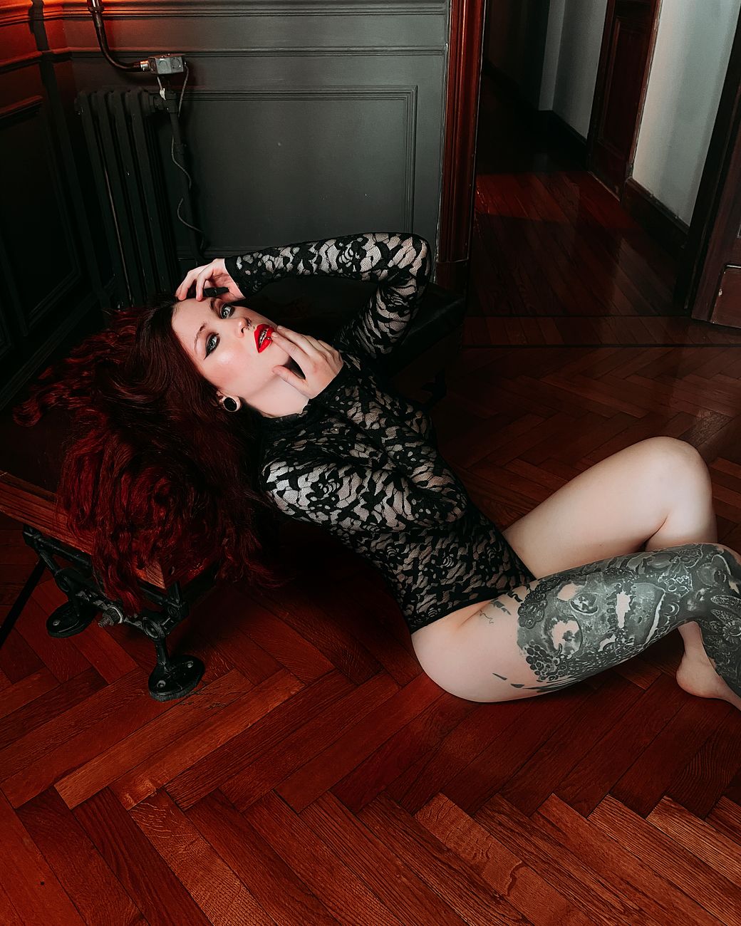 Red-haired woman in lace bodysuit posing sensually indoors on wooden flooring.