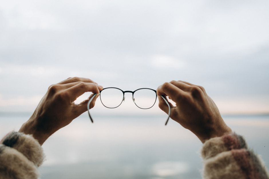 Point of view image of hands holding eyeglasses against a calm lake backdrop.