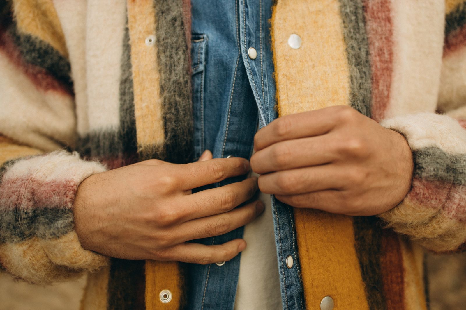Hands buttoning a denim shirt with a striped wool coat — small details like buttons and lining matter more than brand logos.