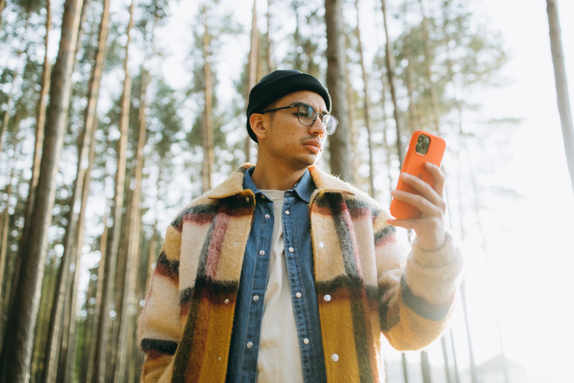 A trendy man in a beanie and plaid coat checks his smartphone amidst tall forest trees.