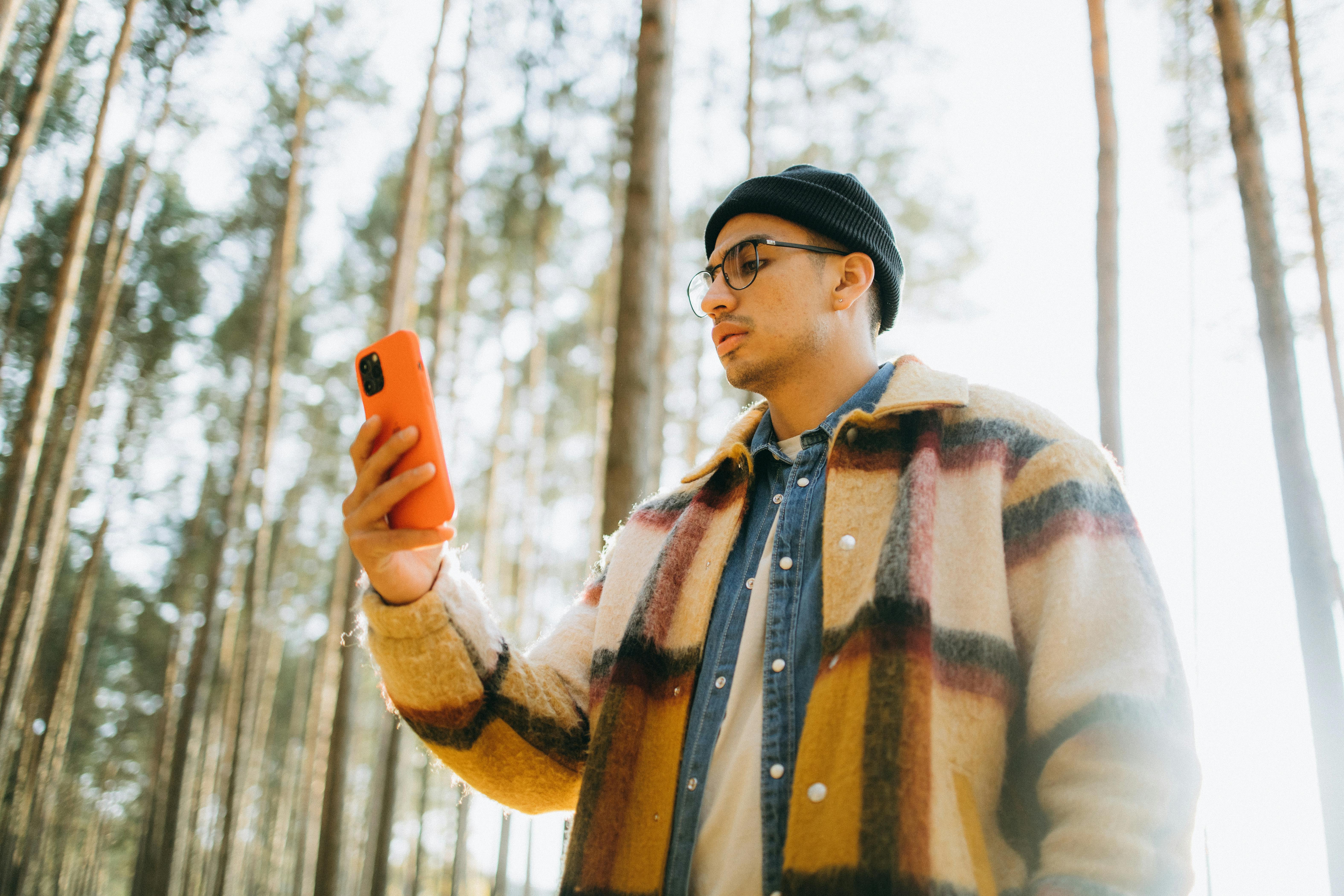 Young man using smartphone in forest, wearing beanie and plaid jacket, low angle view.