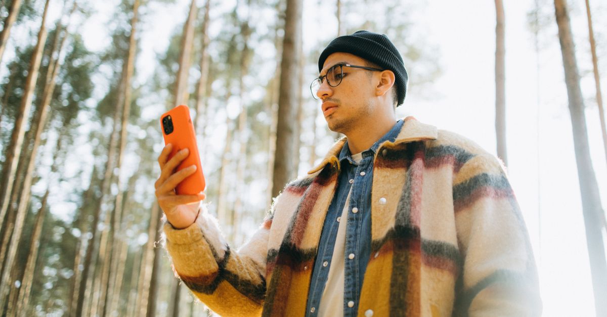 Jovem usando smartphone na floresta, vestindo touca e jaqueta xadrez, vista de baixo.