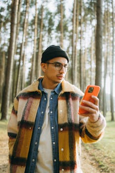 A trendy man in a checkered coat using a smartphone in a serene forest setting.