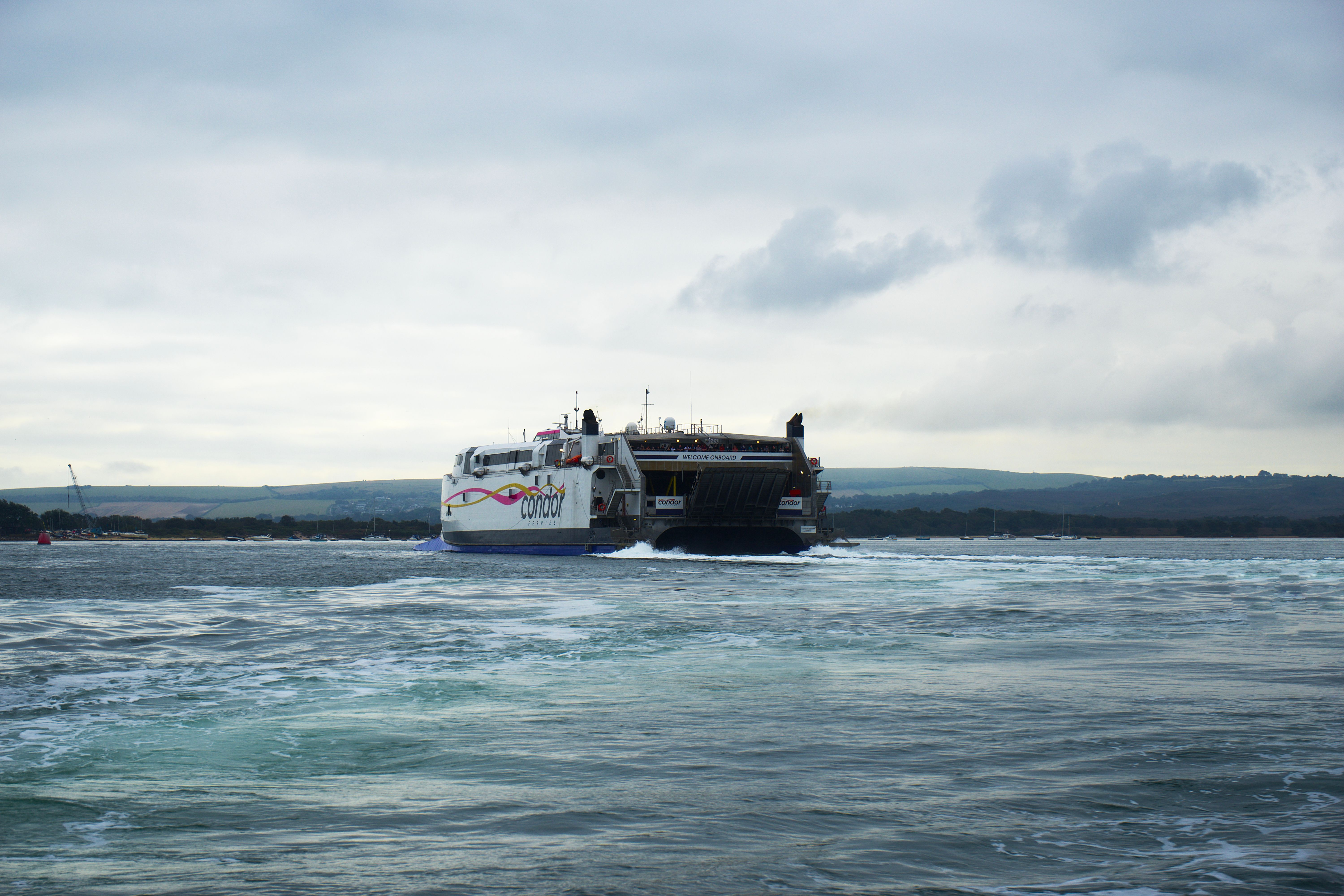 Condor ferry travels across choppy water beneath a cloudy sky, with a low coastline in the distance