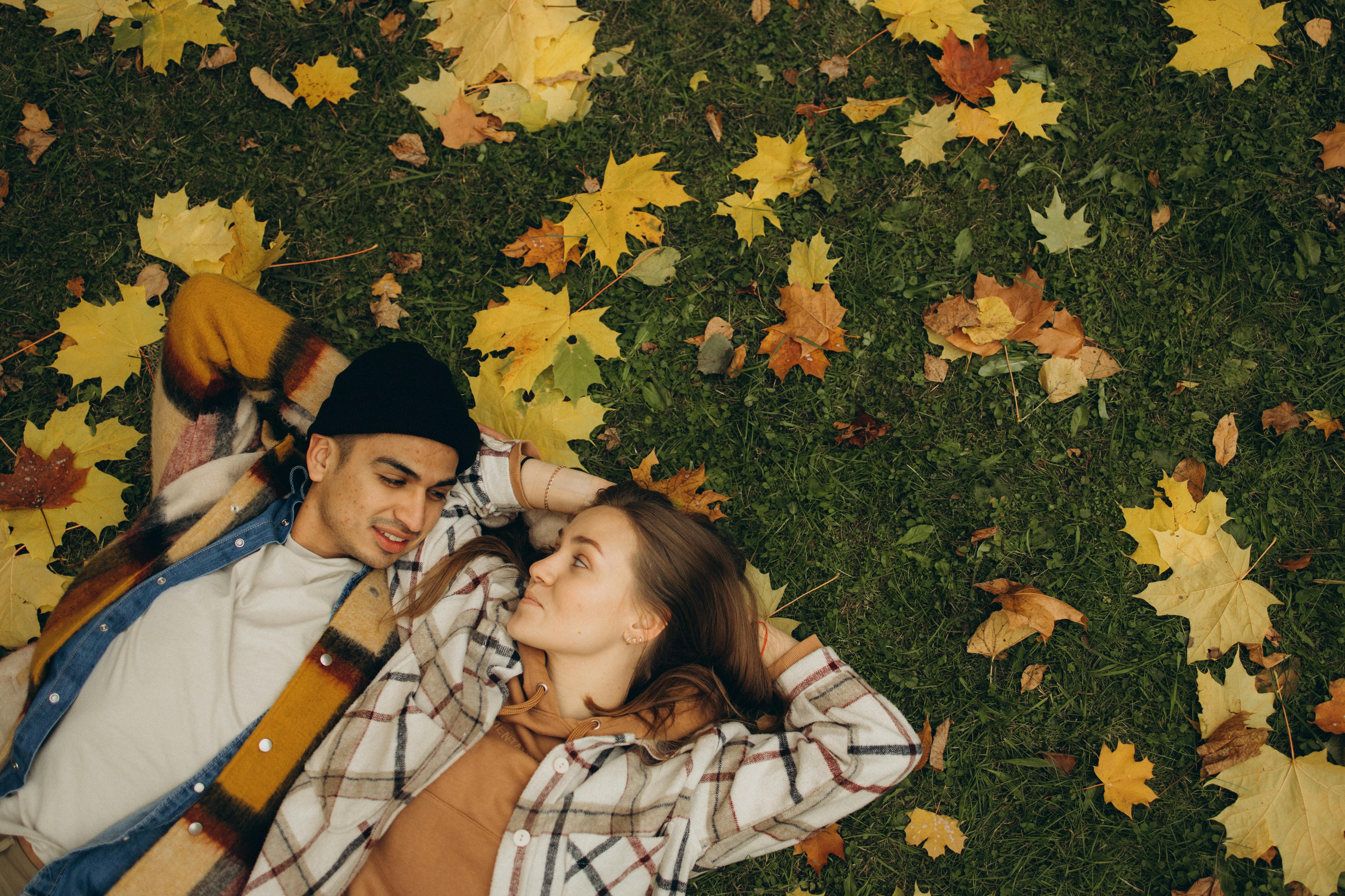 Young couple lying on grass surrounded by autumn maple leaves, embracing seasonal warmth.