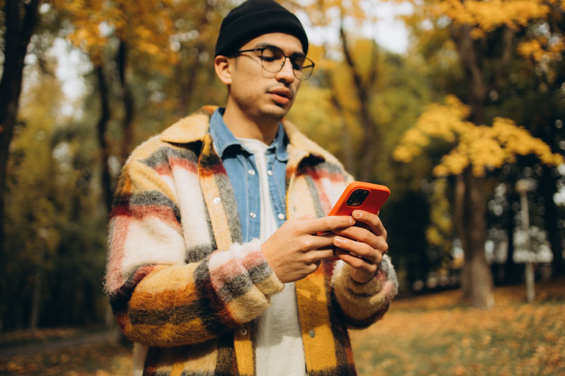 Joven con chaqueta de cuadros usando un smartphone al aire libre en un parque en otoño, concentrándose en la actividad móvil.