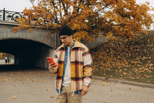 Stylish man using smartphone in park with autumn leaves and vibrant outfit.